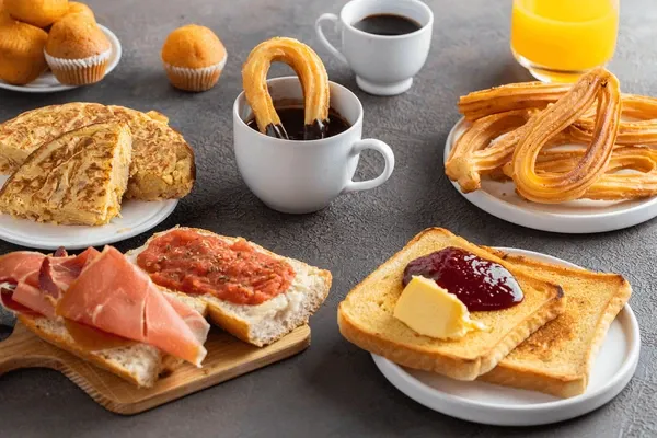 A breakfast table: churros with a cup of thick chocolate, a potato omelette, toast with grated tomato and cured ham, toast with butter and jam, small muffins, coffee and orange juice