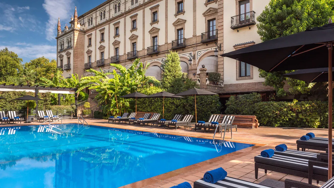 The outdoor pool in the hotel garden, with striped loungers and black parasols along the near side and the hotel's tiled façade and balconies behind banana palms