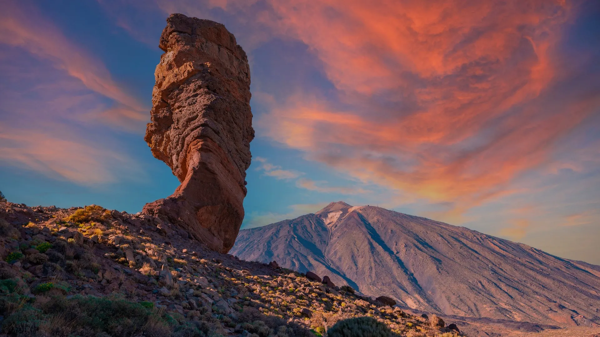 The weathered rock pillar of Roque Cinchado on a stony slope, with the cone of Teide behind it under a pink and orange sky