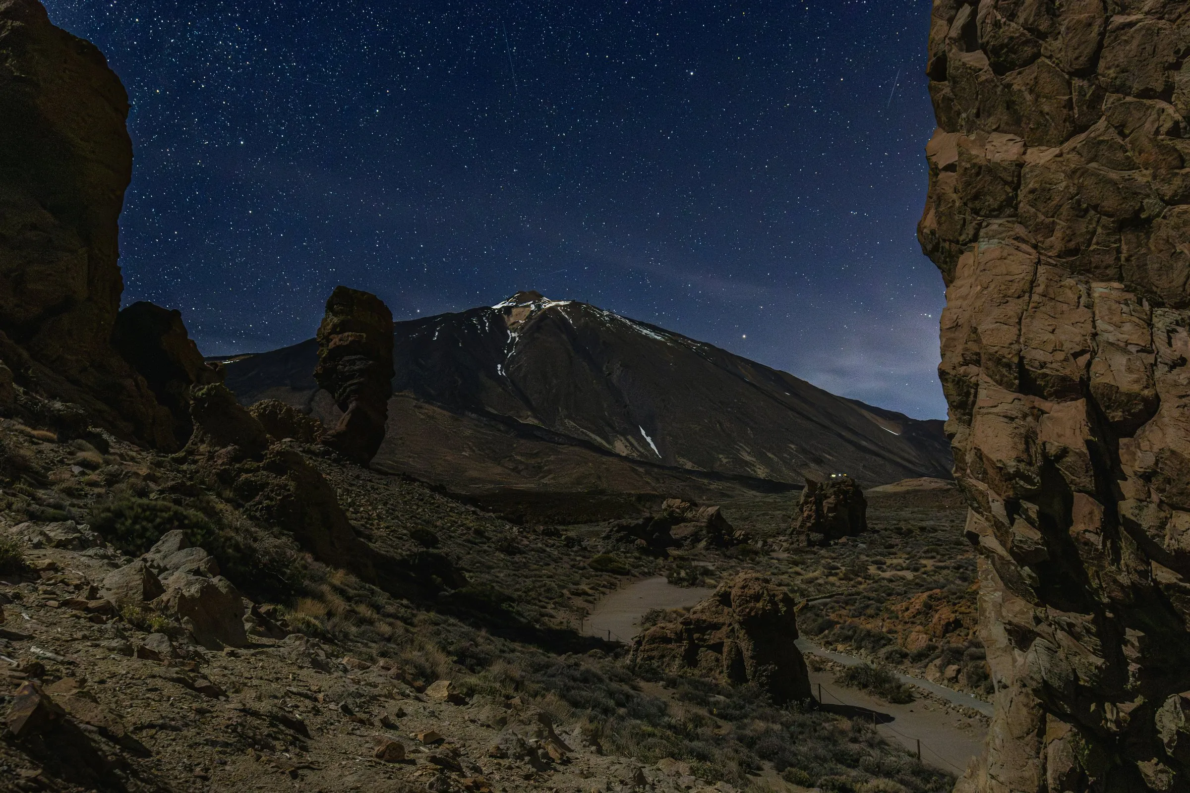 Mount Teide, streaked with snow, under a sky full of stars between dark rock towers of the Roques de García, a footpath winding across the moonlit caldera below
