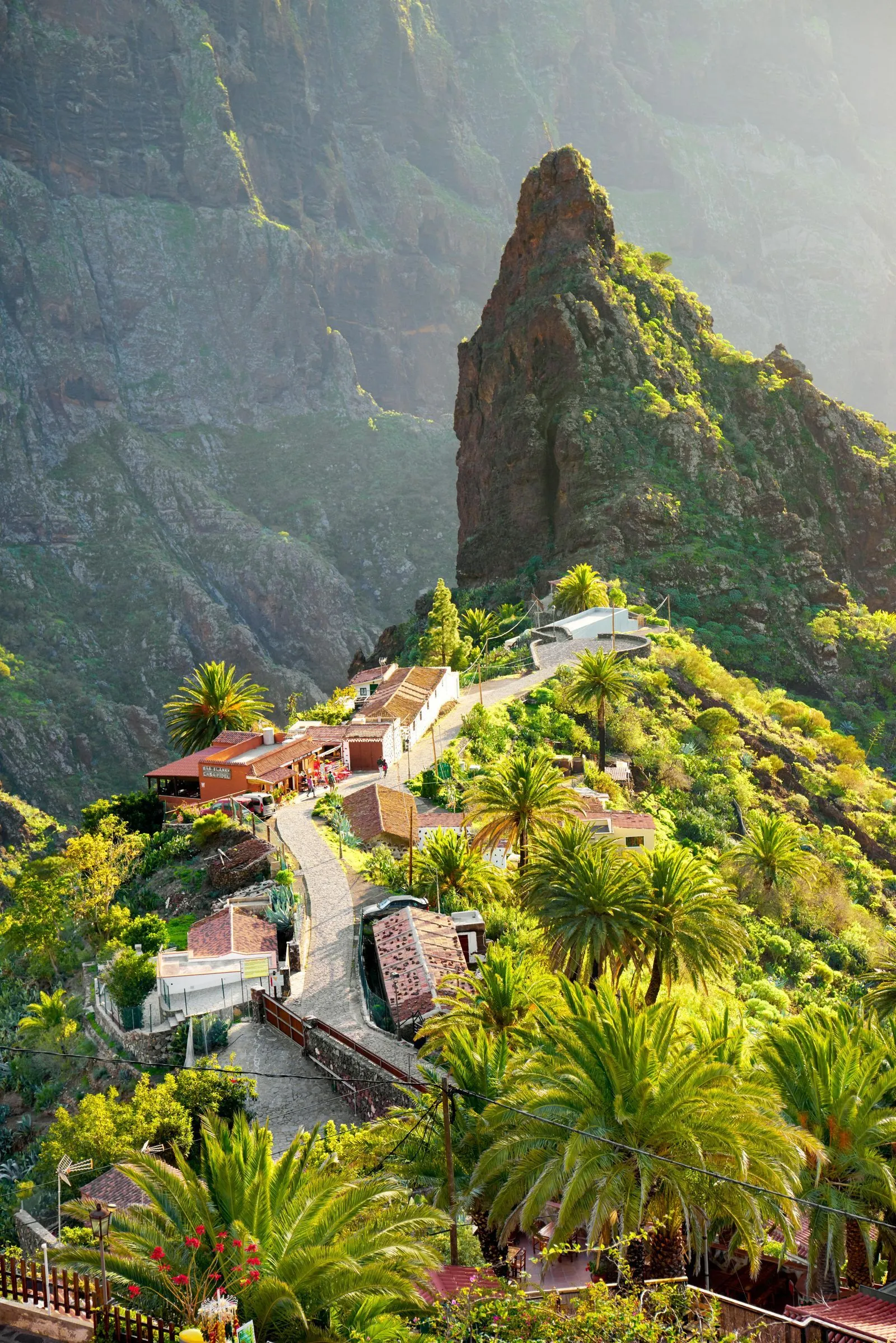 The hamlet of Masca on a green ridge below a tall rock pinnacle, palms and terraces along a narrow lane, the walls of the gorge behind