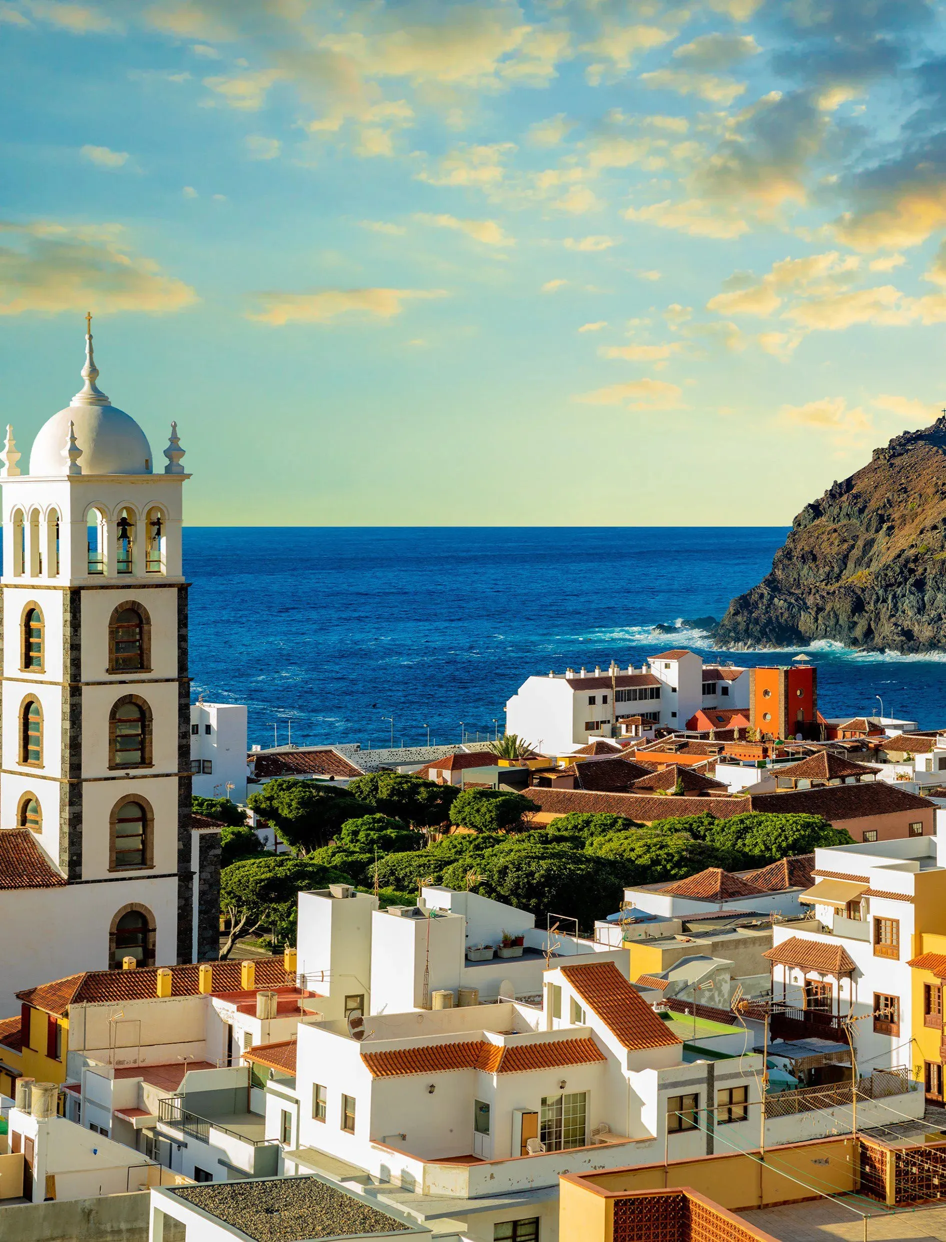 Garachico from above at golden hour: a white bell tower with a domed top, red-tiled roofs, the Atlantic and a dark headland behind