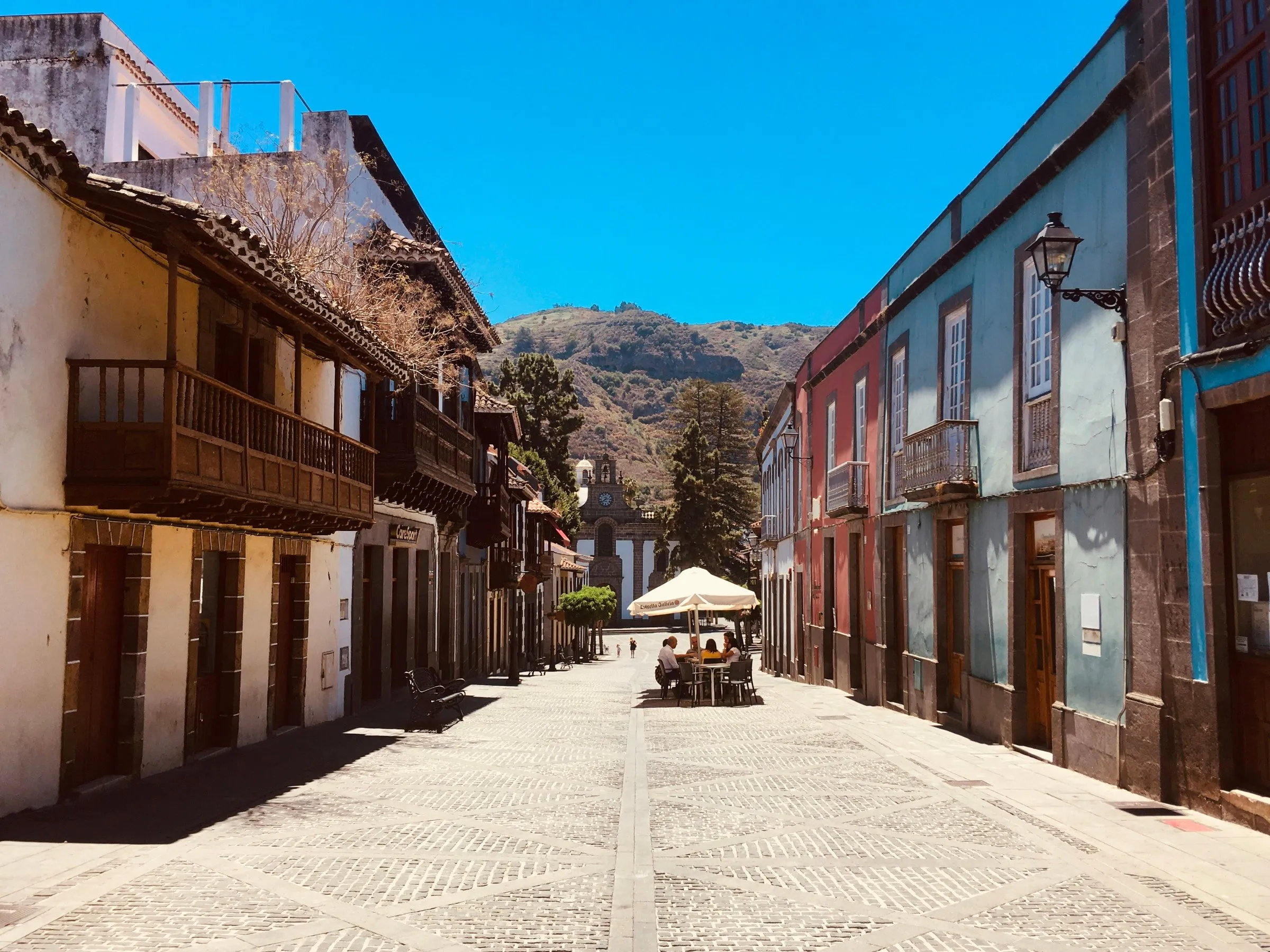 A paved street in Teror between old houses with dark carved wooden balconies, a café parasol halfway down and the basilica's tower at the end, a green hill behind