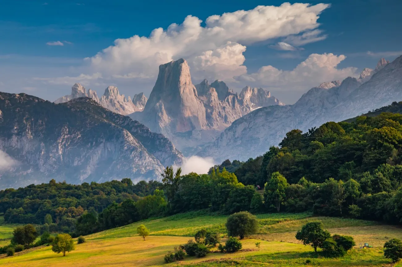 The pale limestone tower of the Naranjo de Bulnes rising among grey peaks above green meadows and woods, under tall white summer clouds