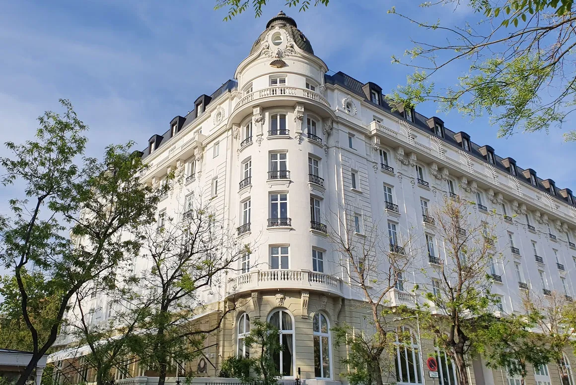 The white corner tower of the Mandarin Oriental Ritz in Madrid, rising past balconied floors to a slate dome, among spring trees under a blue sky