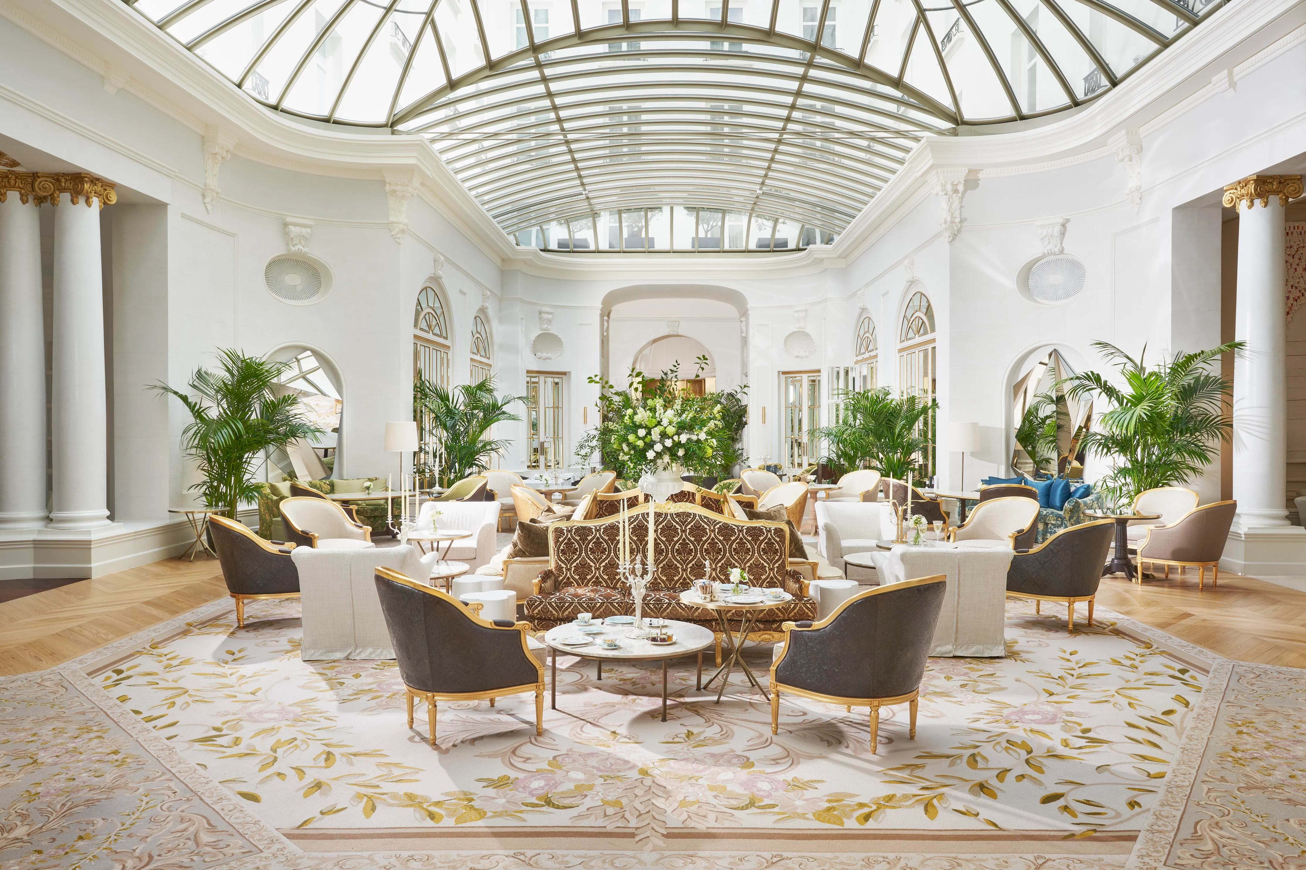 A court under an arched glass roof, with white walls, columns and arched doorways, palms and a tall arrangement of white flowers, and gilded armchairs and sofas
