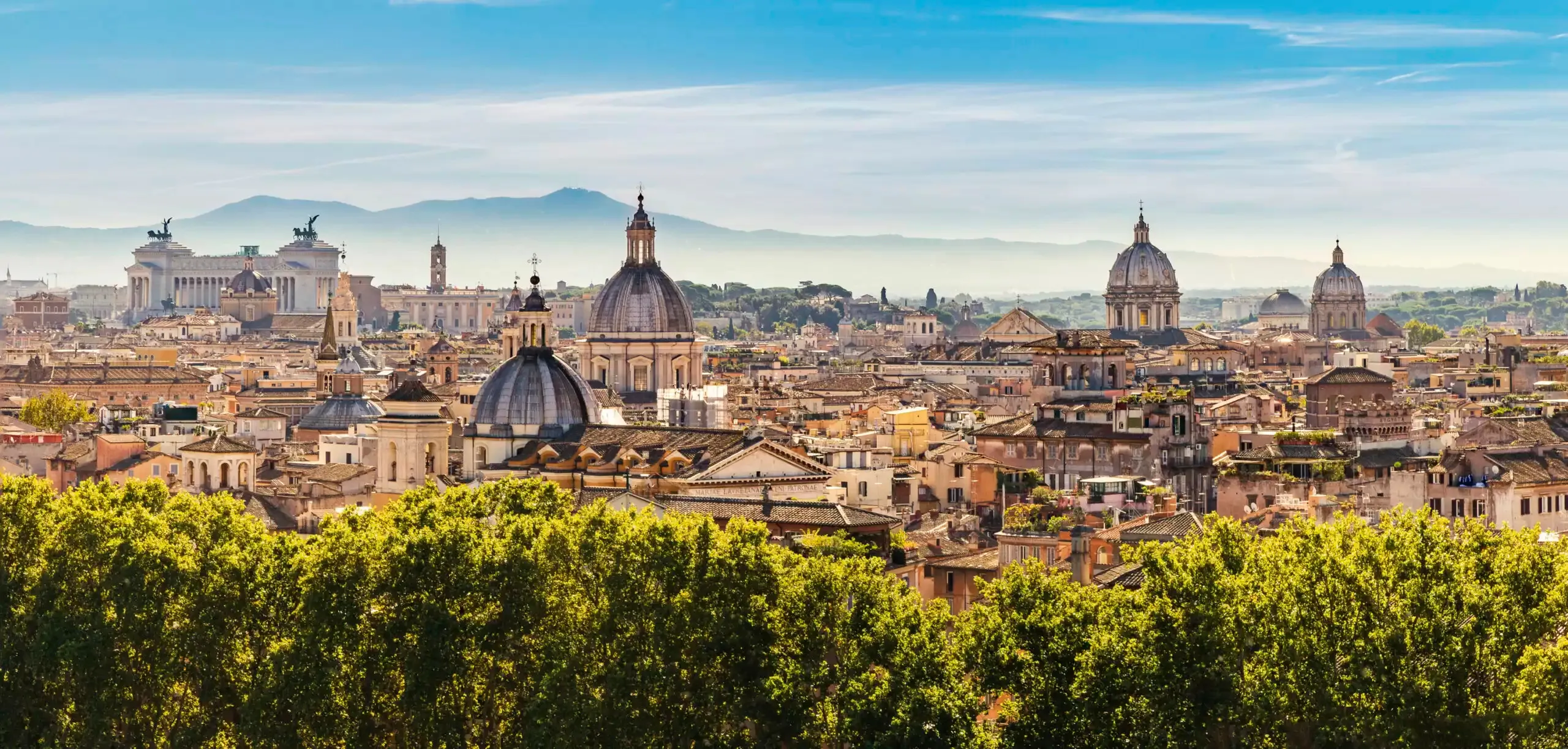 Rome's rooftops and domes in bright sun above a line of trees, the white colonnade of the Vittoriano with its two bronze chariots on the left, a tall grey dome at the centre, more domes to the right and blue hills along the horizon