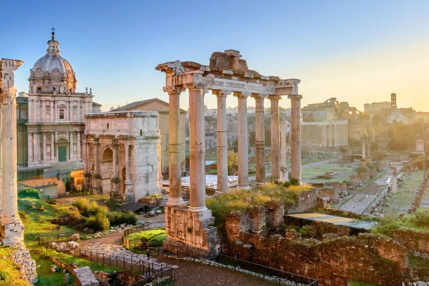 The columns of the Temple of Saturn in the Roman Forum lit by low morning sun, a triumphal arch and a domed Baroque church behind them on the left, and the ruins of the Forum running away to the right