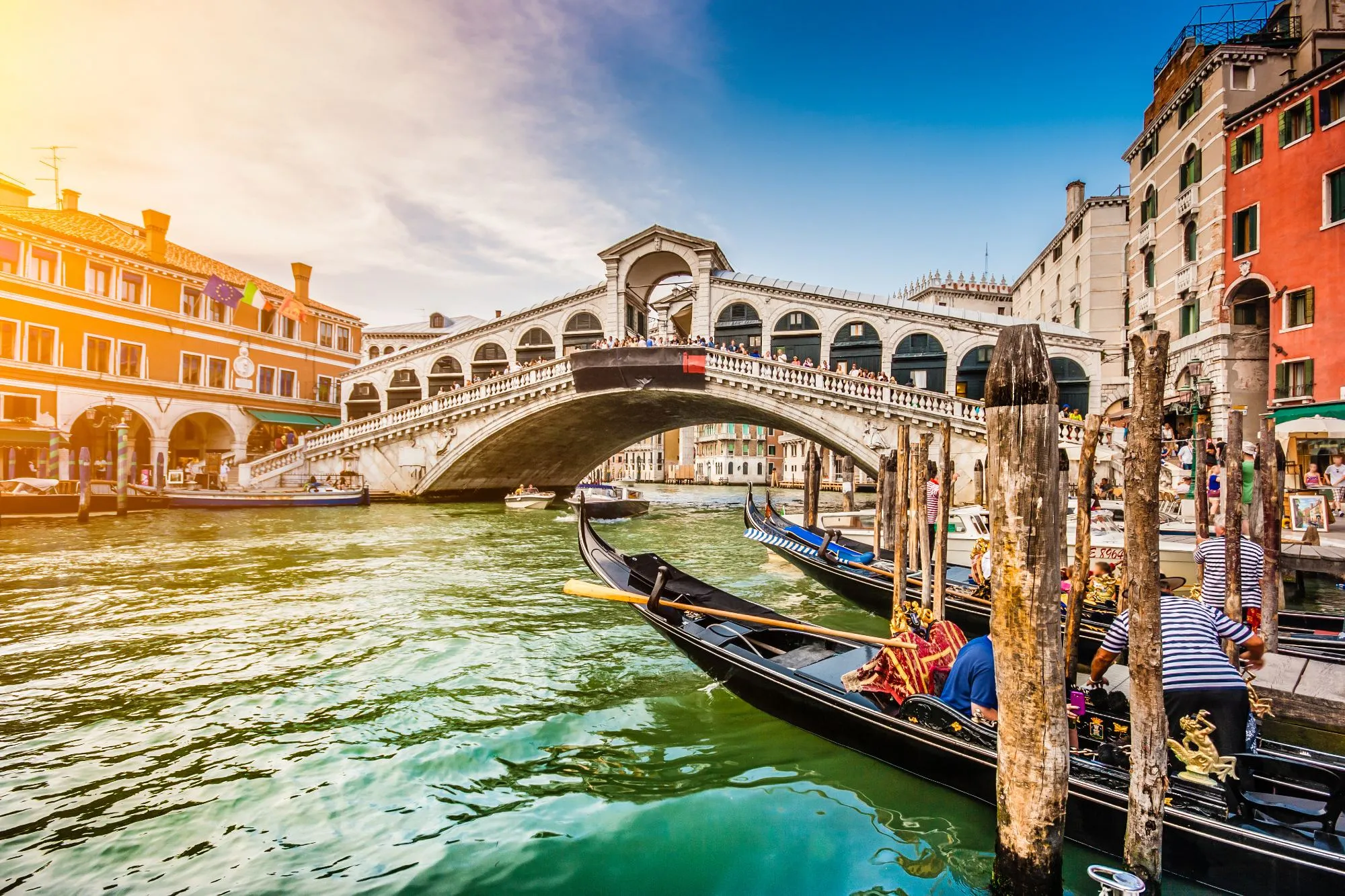 The Rialto Bridge over the Grand Canal in late-afternoon sun, gondolas moored between wooden posts in the foreground with a gondolier beside them, and painted palazzi lining the water on either side