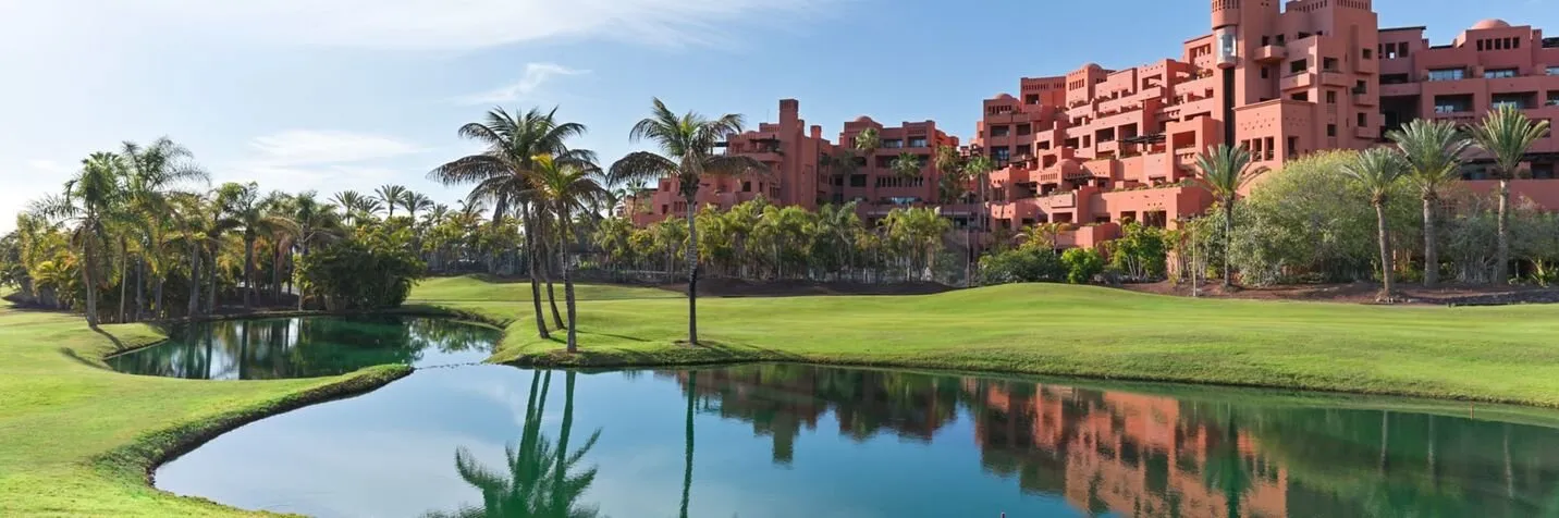 The resort's terracotta blocks rising behind palms above a fairway, with a still lake in the foreground reflecting the building