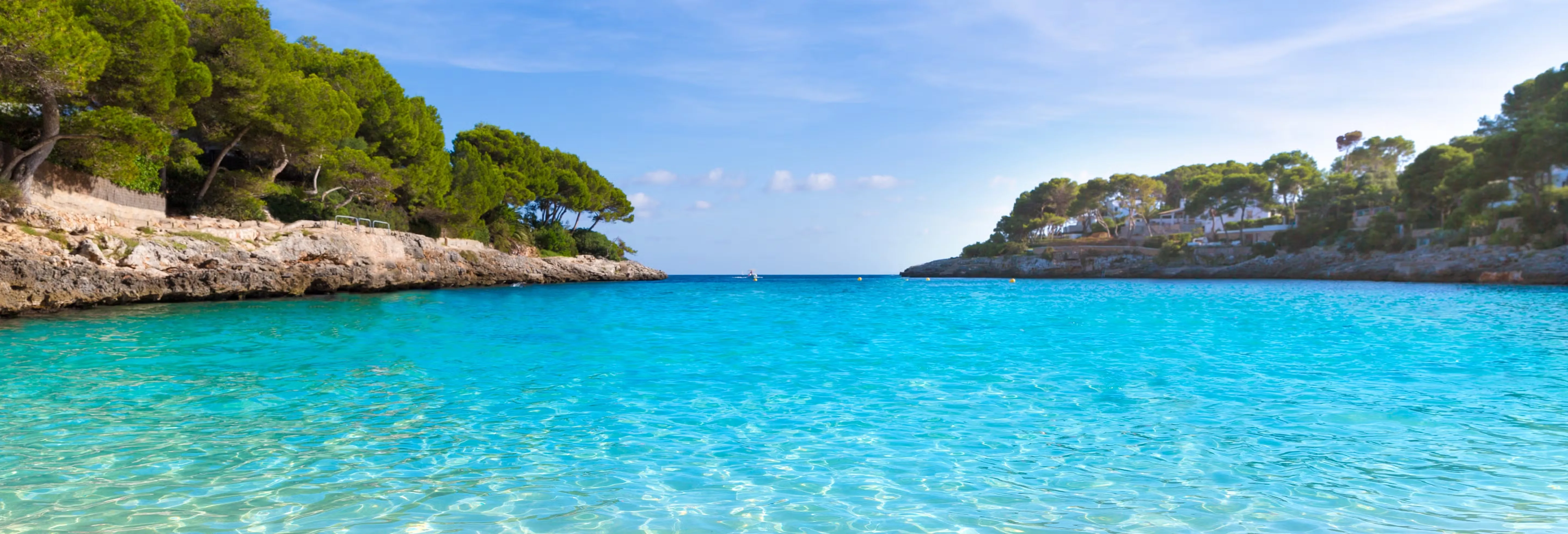 A cove of clear turquoise water between low rocky shores shaded by pine trees, houses among the pines on the right and the open sea beyond the mouth of the inlet