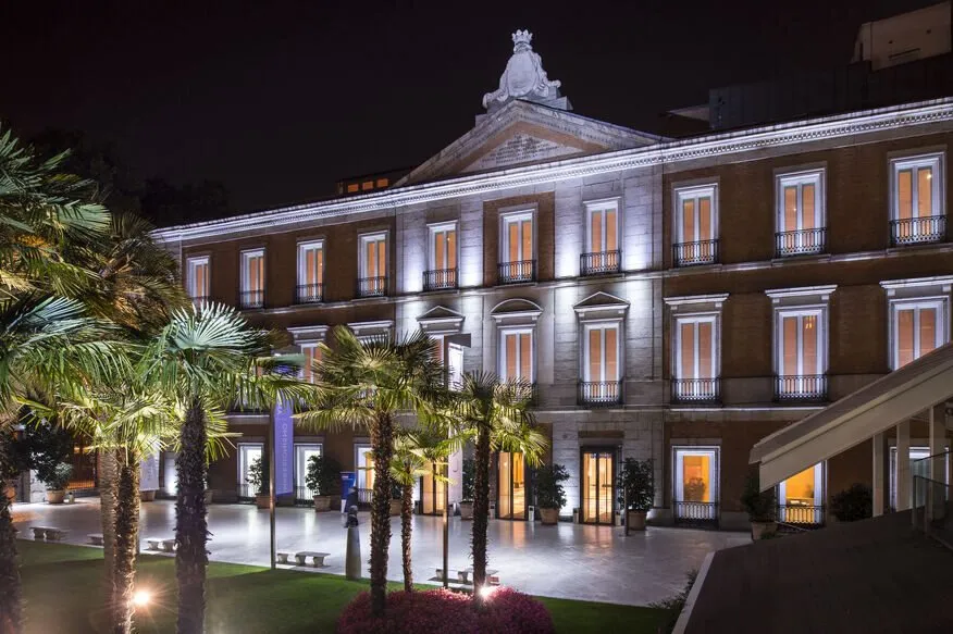 The Thyssen-Bornemisza museum at night: a long brick and stone palace front with tall lit windows and iron balconies, a sculpted crest on the pediment, and floodlit palms in the garden before it
