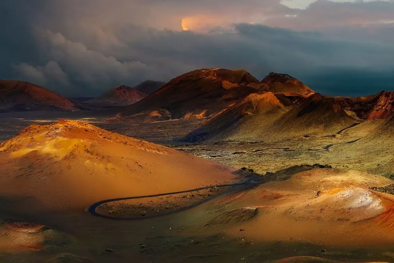 Red and ochre volcanic cones under a dark, stormy sky, lit by a break of low sun, with a narrow road looping between them across the dark lava
