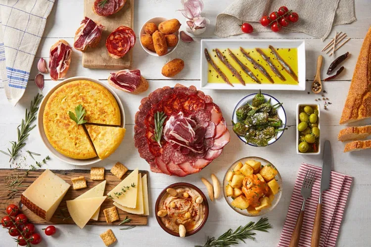 A spread of Spanish tapas from above on a white table: a potato omelette, a platter of chorizo and cured ham, anchovies in olive oil, blistered green peppers, croquetas, fried potatoes in sauce, wedges of cheese and bread