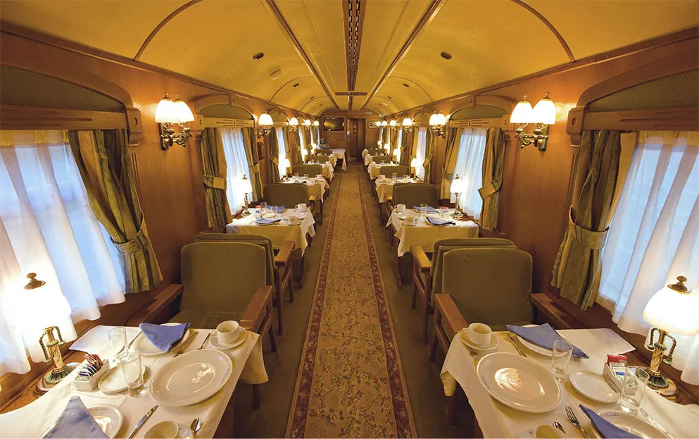 A railway dining car with tables laid in white for breakfast, green upholstered armchairs, brass lamps on the tables, curtained windows and a patterned runner down the aisle under a curved wooden ceiling