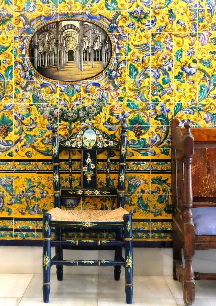 A dark blue chair painted with flowers, with a rush seat, against a wall of yellow and blue tiles whose inset panel shows the striped arches of a mosque; a worn wooden settle beside it