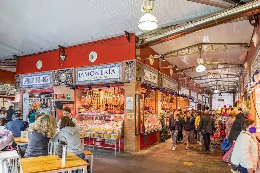 Inside a covered market: tiled stall fronts for spices, a jamonería hung with hams and a cheese stall, with shoppers in the aisle