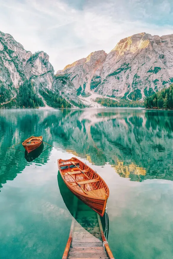 Two wooden rowing boats on the still green water of Lago di Braies, a wooden jetty in the foreground, and the pale cliffs and pine slopes of the Dolomites reflected behind