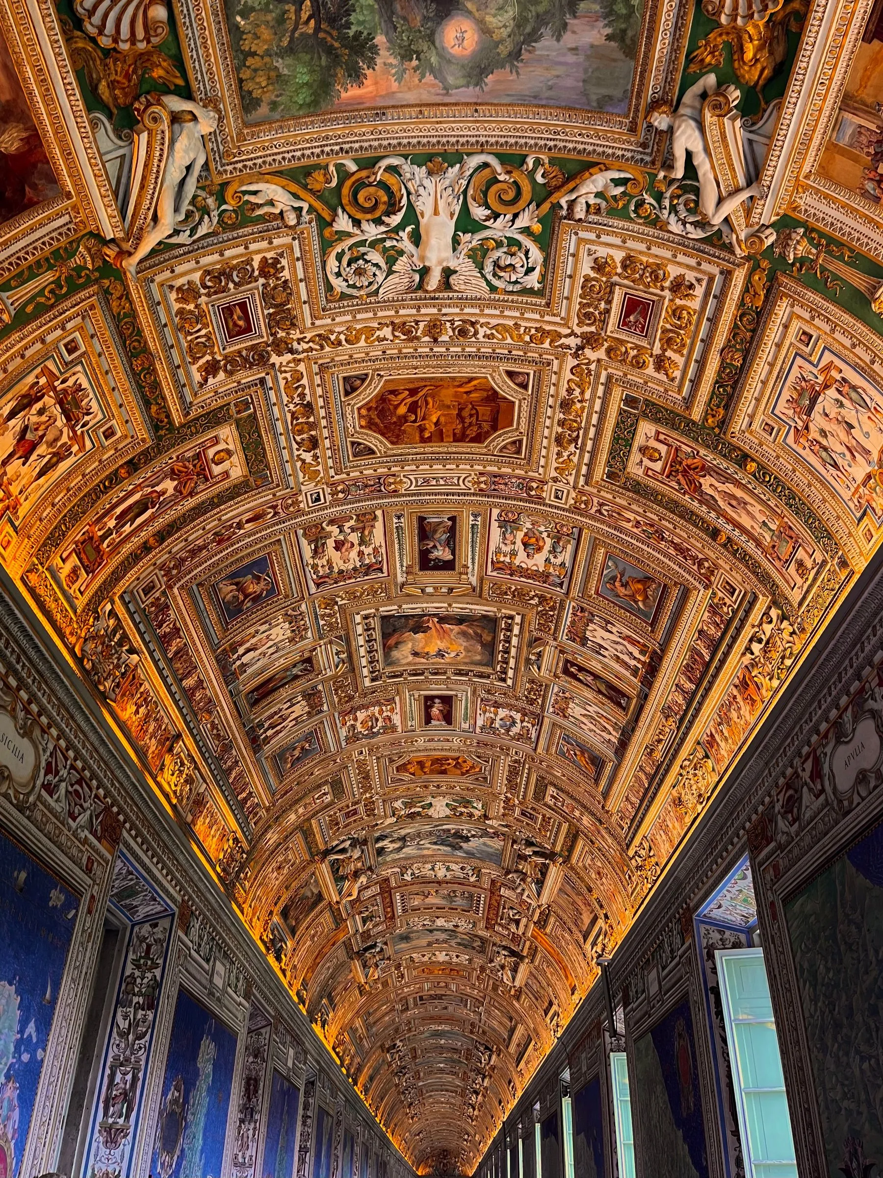 The Gallery of Maps in the Vatican Museums seen along its length from below: the long barrel vault covered in gilded stucco and painted panels running away to a far door, with the blue and green painted maps of Italy on the walls at either side