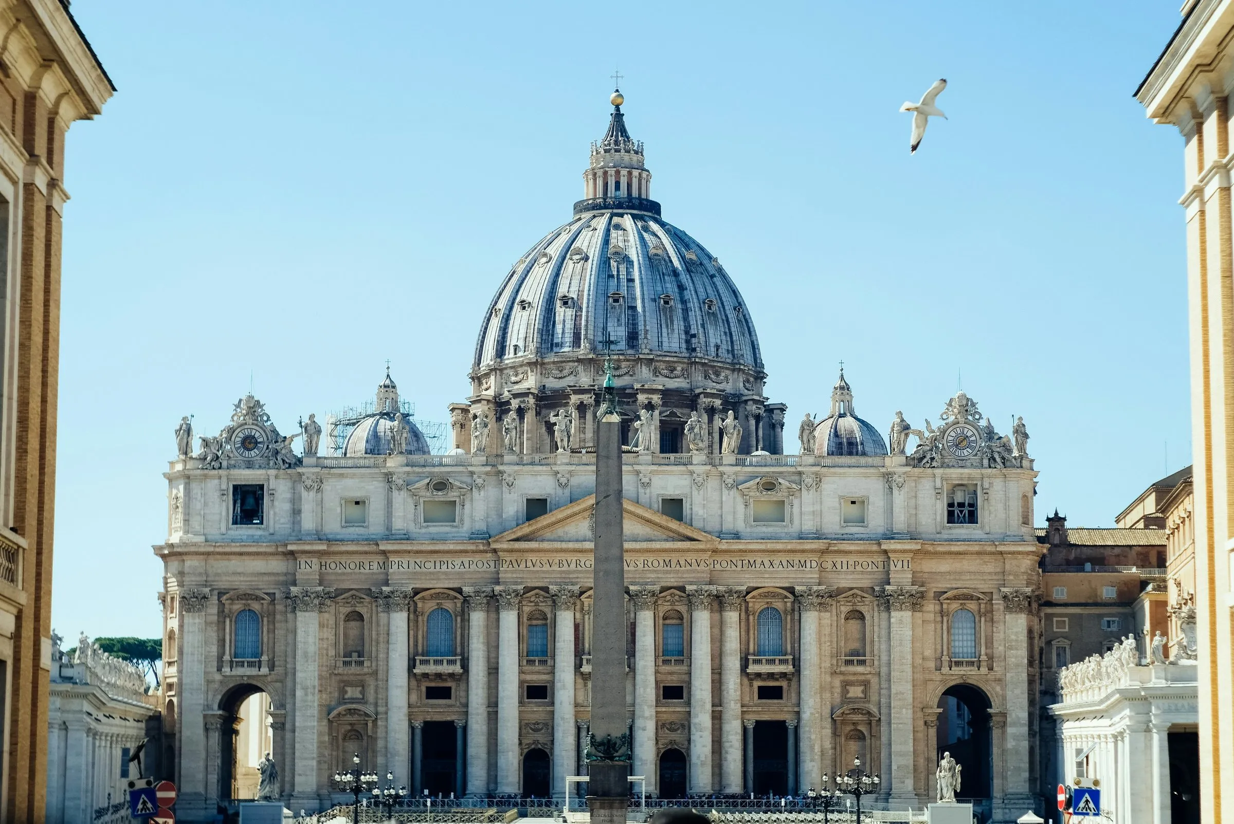 St Peter's Basilica straight on: Michelangelo's dome above Maderno's façade, the obelisk of St Peter's Square in front, buildings framing the view at either side and a gull in a clear blue sky