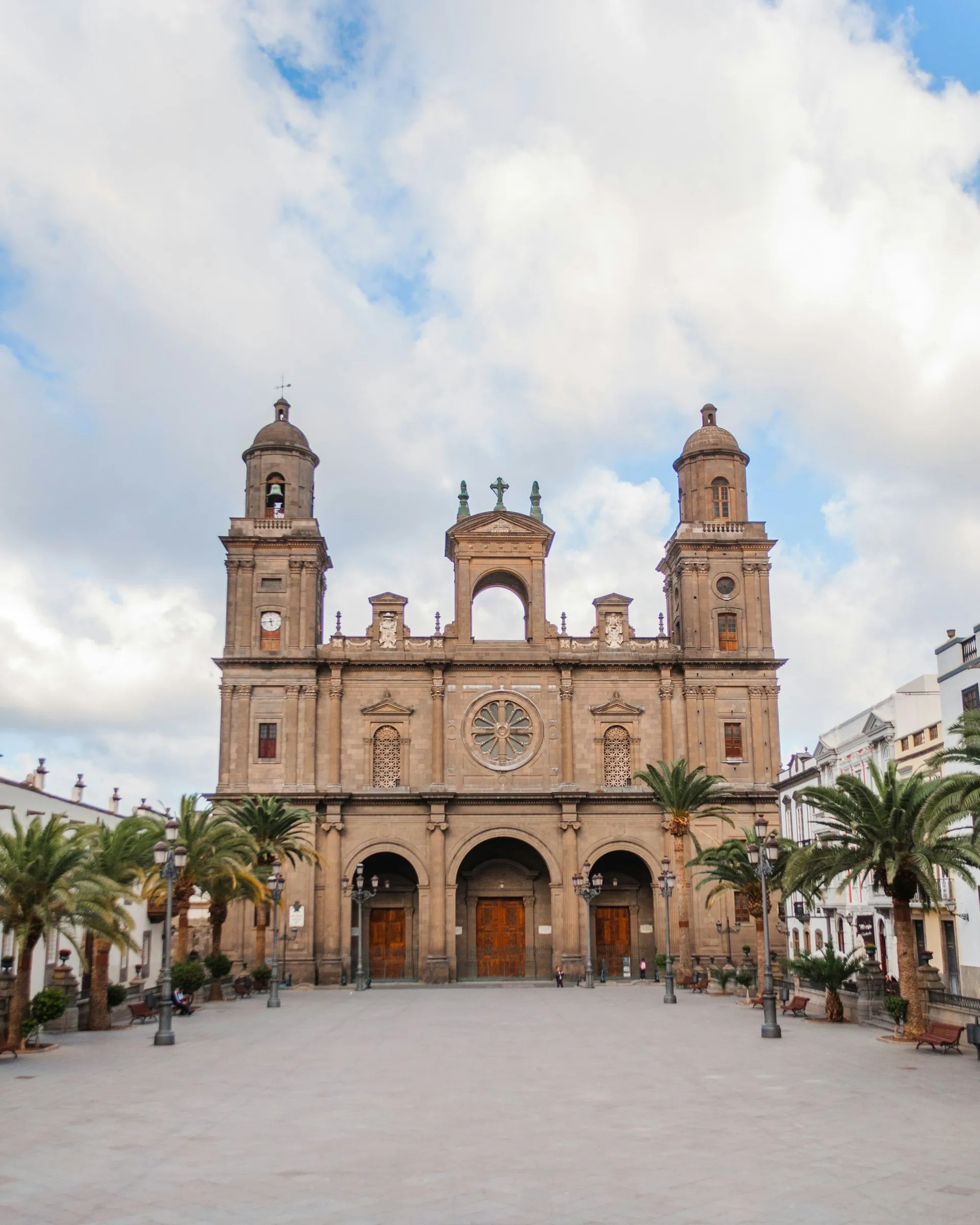 The stone front of the Cathedral of Santa Ana, two domed bell towers and three arched doors, over a wide paved square lined with palms