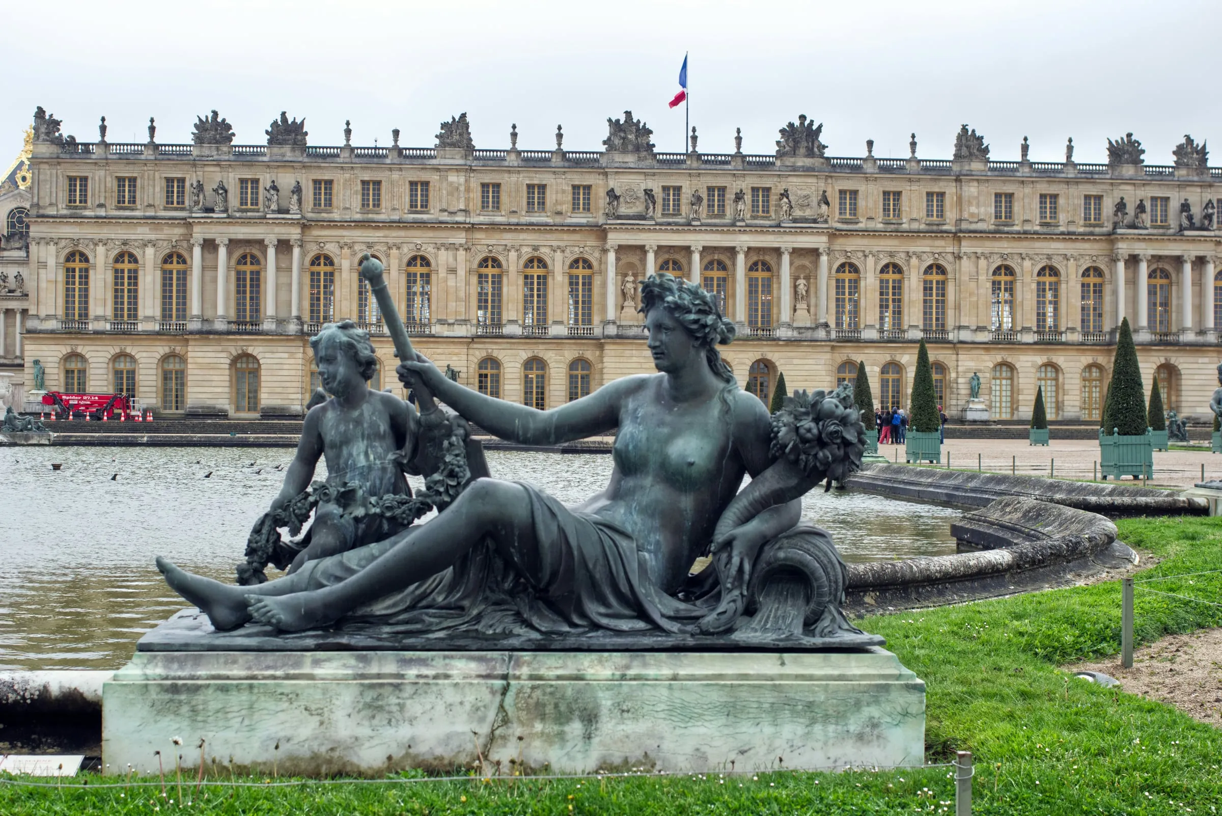 A reclining bronze river figure with a child beside her on the rim of a basin in the gardens of Versailles, the long garden front of the palace behind under a grey sky
