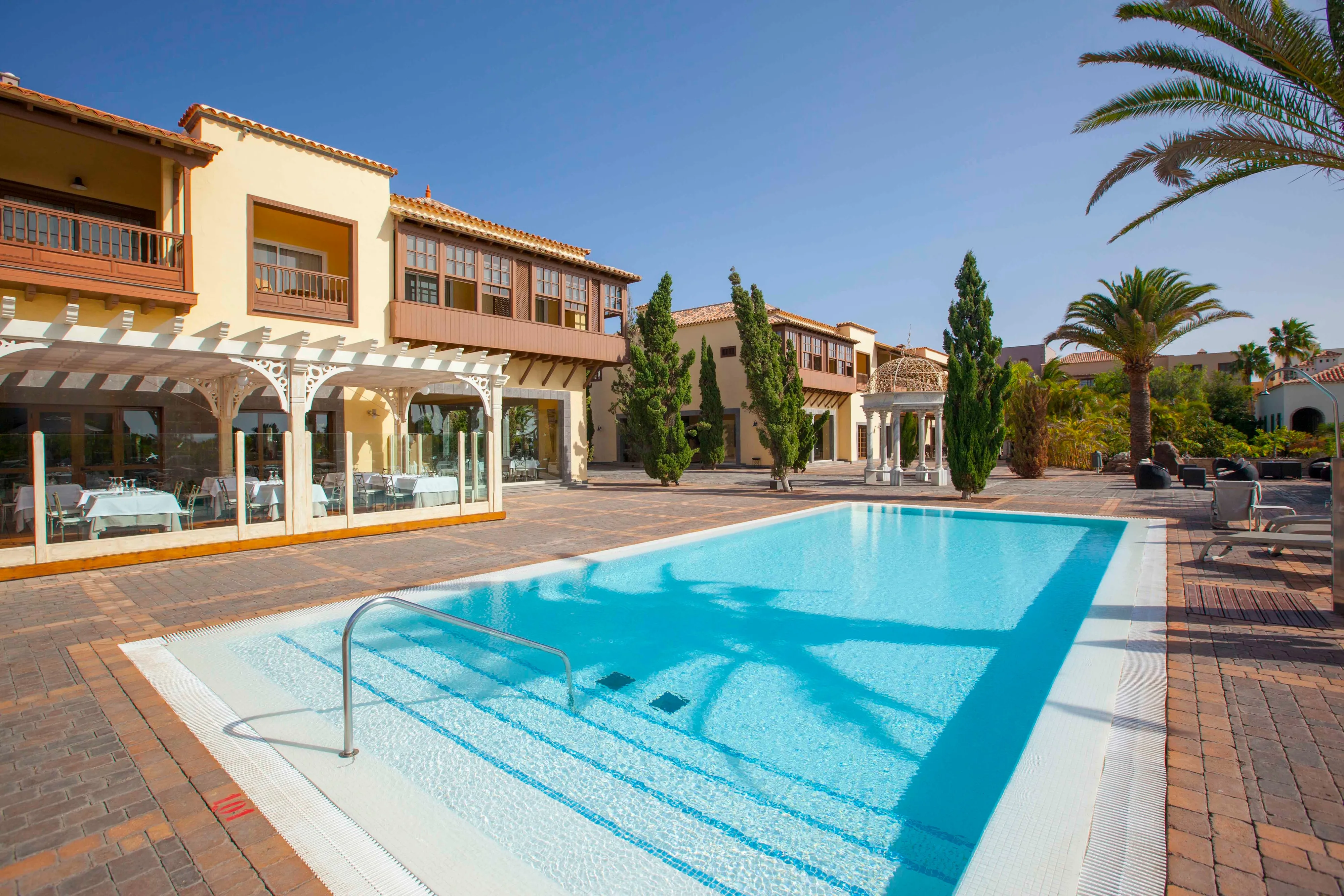 The Arcos pool: a long rectangular pool on terracotta paving, ochre two-storey houses with dark-wood Canarian balconies and a white pergola behind it, cypresses, a white domed gazebo and palms under a clear sky