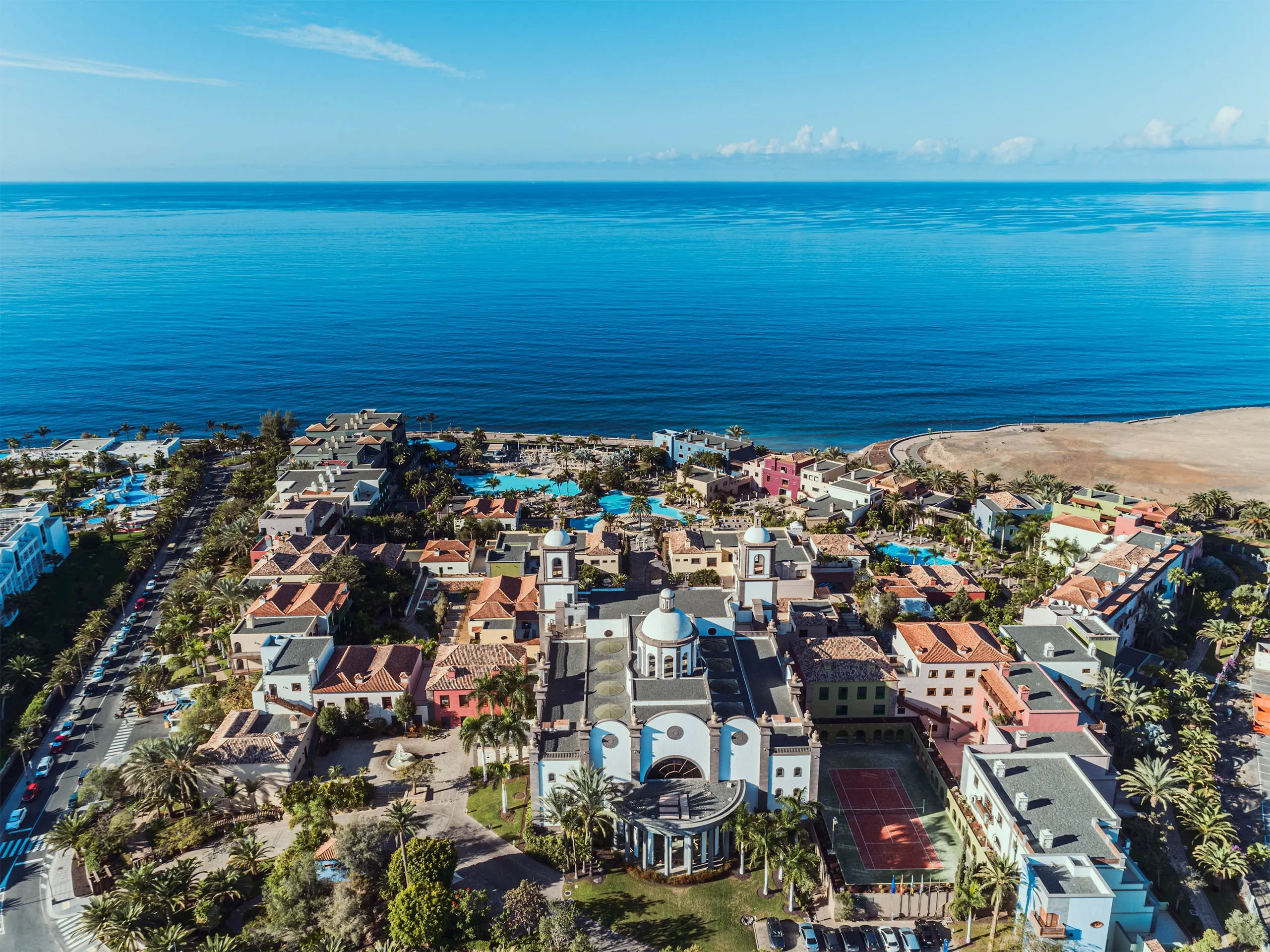 Lopesan Villa del Conde from the air: a white church-like main building with two bell towers and a dome at its centre, low houses with terracotta and grey roofs in cream, pink and green around it, palms, blue pools and a tennis court, the deep-blue Atlantic filling the upper half of the frame