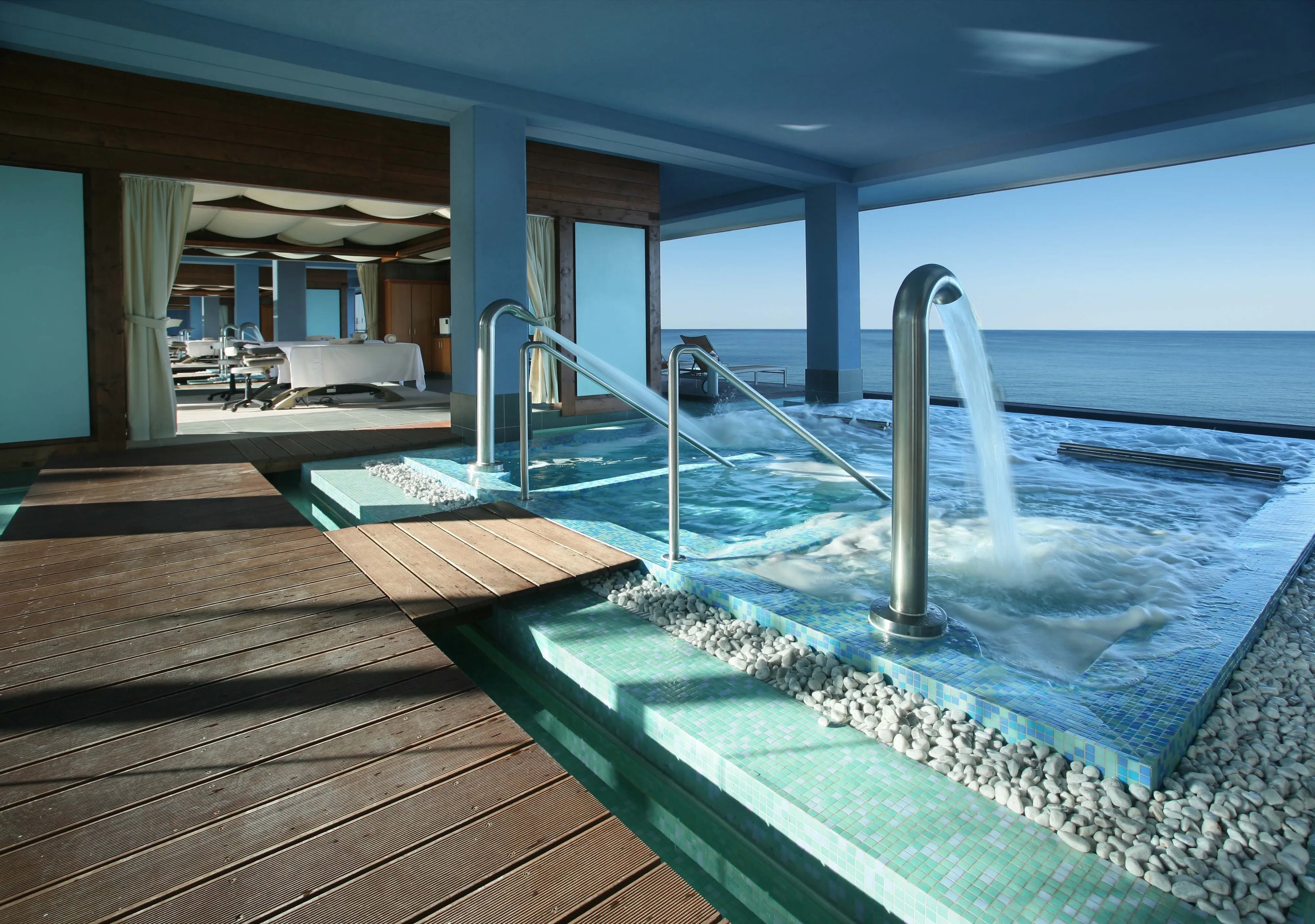 Om Thalasso's open-sided hydrotherapy pool: steel water jets and rails in a mosaic pool edged with white pebbles and a timber deck, treatment beds behind cream curtains at left, the calm sea and a pale sky through the open side