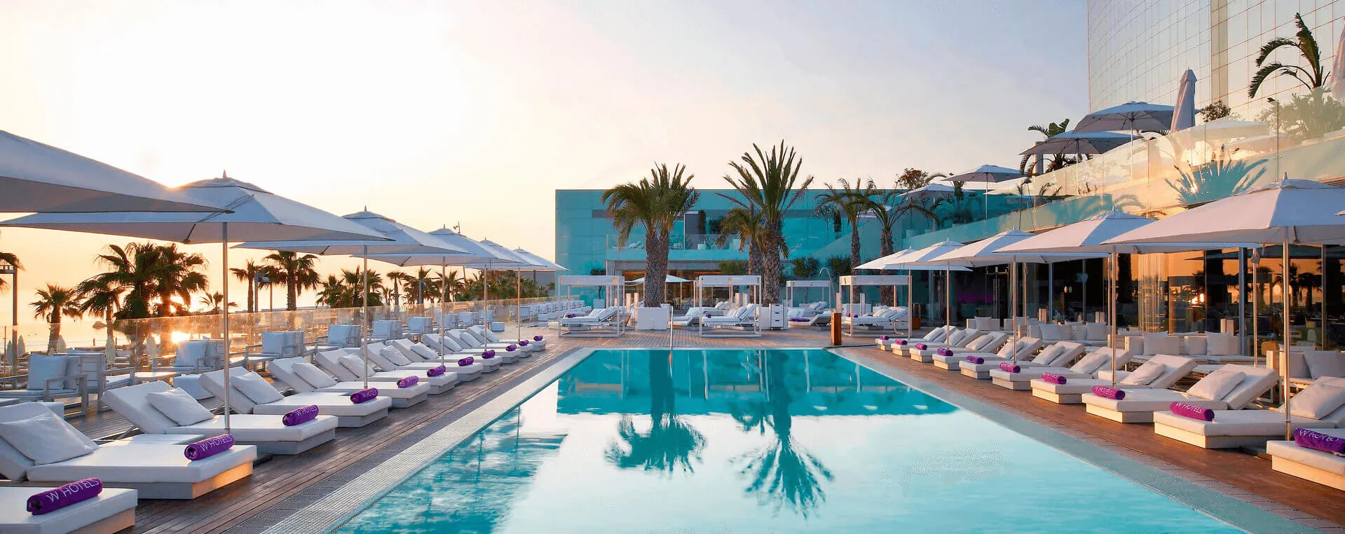 A long turquoise pool at sunset reflecting palm trees, rows of white loungers with purple rolled towels under white parasols on a timber deck, palms along the sea wall on the left and the hotel's glass tower rising on the right